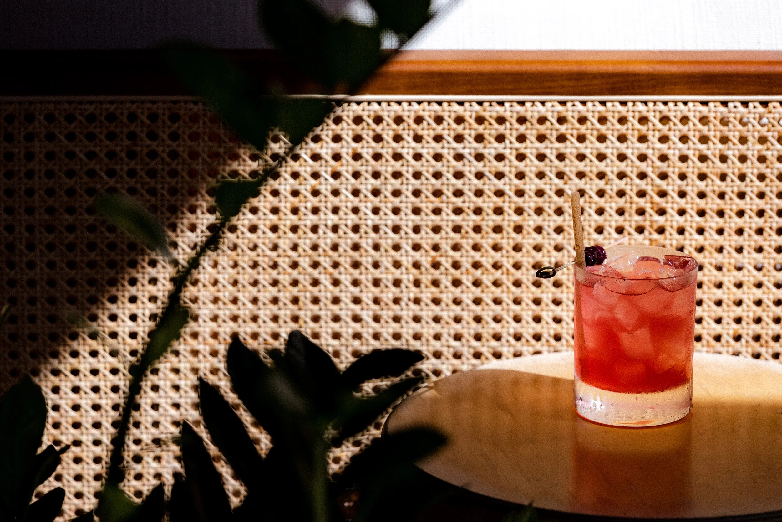 A pink cocktail with a sprig of garnish on a small round table, dappled sunlight falling across a cane-back panel.